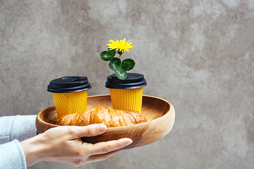 Two yellow paper cups for coffee or tea and Fresh croissant on a wooden tray. Takeaway order in women's hands. Dandelion flower in the lid of a container for a romantic breakfast. Copy space for text