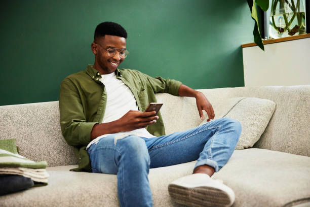 istock Smiling young sitting on a sofa and texting on his mobile phone 1407081458