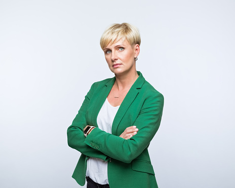 Portrait of mature woman wearing green jacket, standing with arms crossed and looking at camera. Studio shot, grey background.