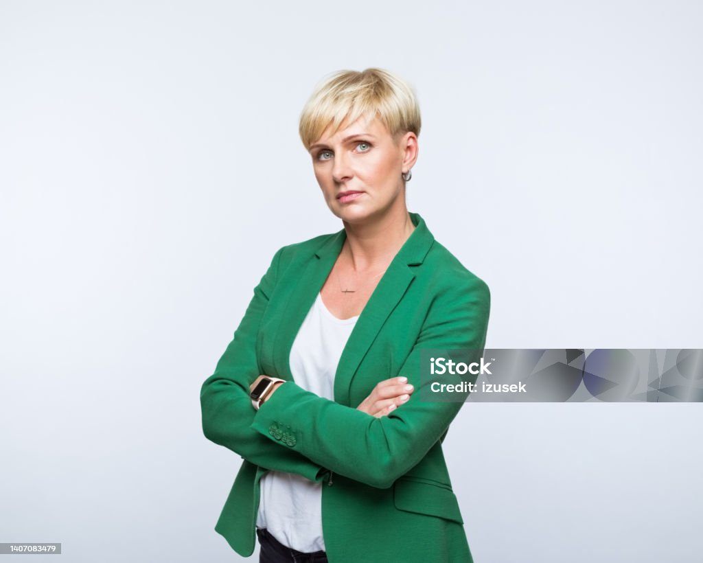 Portrait of confident businesswoman Portrait of mature woman wearing green jacket, standing with arms crossed and looking at camera. Studio shot, grey background. Serious Stock Photo