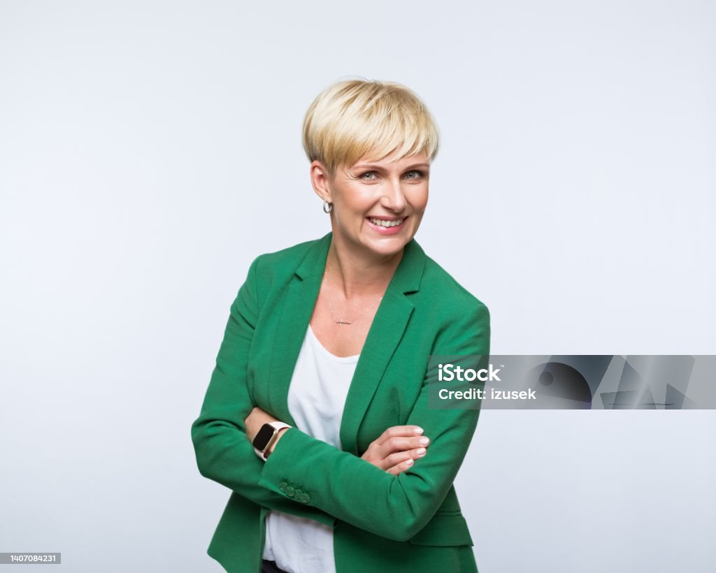 Portrait of happy businesswoman Portrait of confident mature woman wearing green jacket, standing with arms crossed and smiling at camera. Studio shot, grey background. Businesswoman Stock Photo