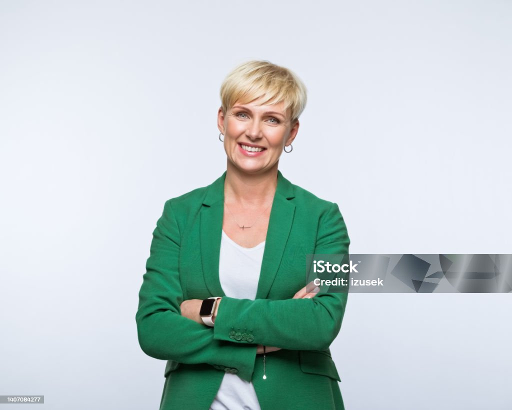 Portrait of happy businesswoman Portrait of confident mature woman wearing green jacket, standing with arms crossed and smiling at camera. Studio shot, grey background. Mature Women Stock Photo