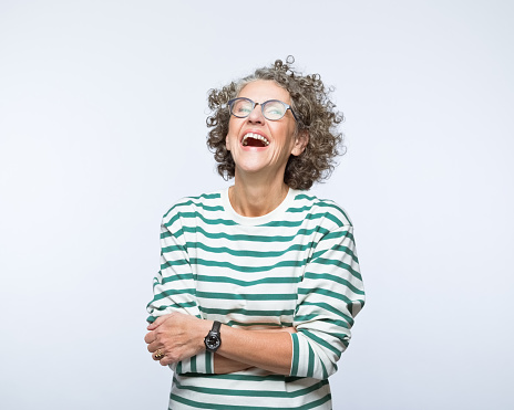 Portrait of confident mature woman wearing striped blouse and eyeglasses, standing with arm crossed and laughing. Studio shot, grey background.