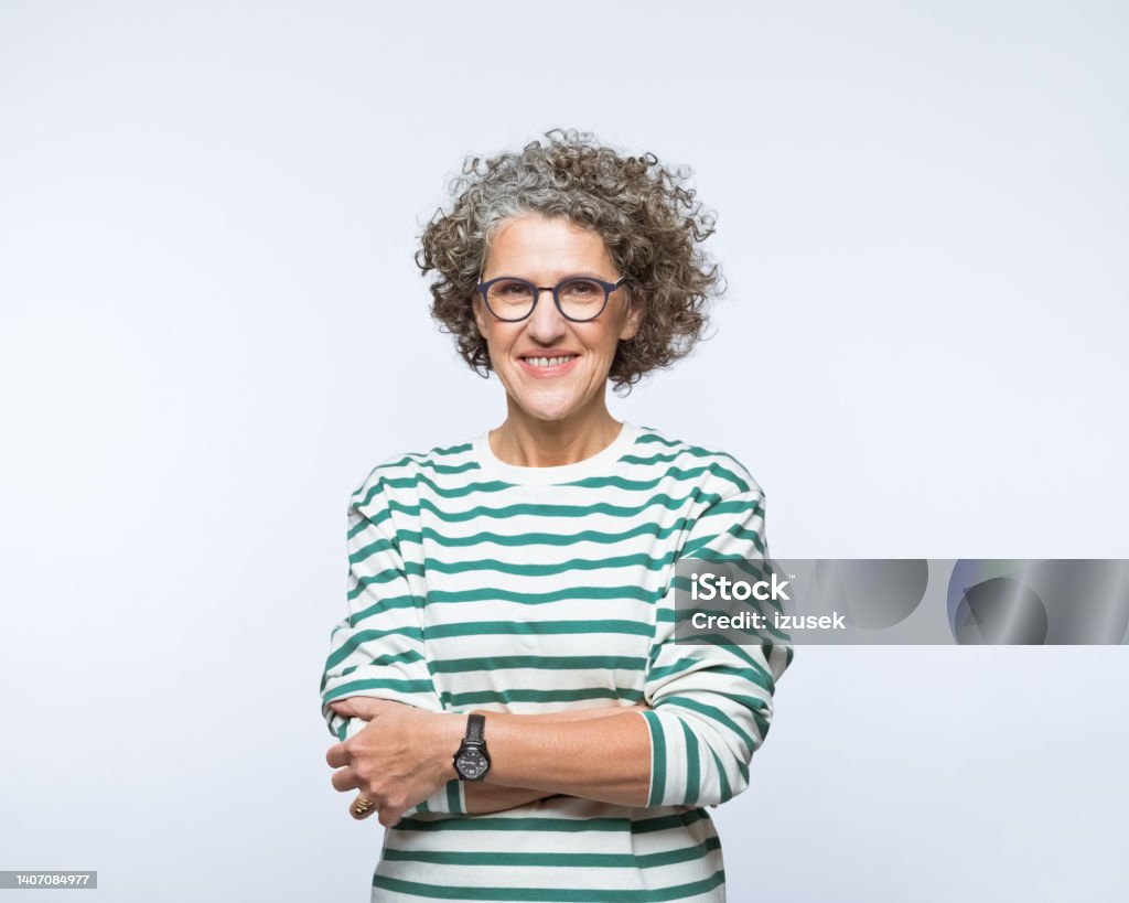 Portrait of smiling mature women Portrait of confident mature woman wearing striped blouse and eyeglasses, standing with arm crossed and smiling at camera. Studio shot, grey background. Arms Crossed Stock Photo