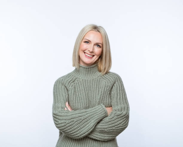 Portrait of smiling mature women Portrait of confident mature woman wearing khaki sweater, standing with arms crossed and smiling at camera. Studio shot, grey background. older blonde woman stock pictures, royalty-free photos & images