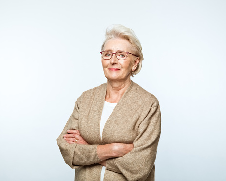 Portrait of confident senior woman wearing beige cardigan, standing with arms crossed and smiling at camera.. Studio shot, grey background.