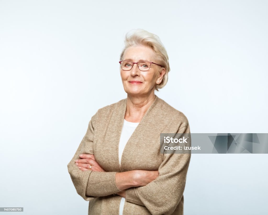 Portrait of smiling senior women Portrait of confident senior woman wearing beige cardigan, standing with arms crossed and smiling at camera.. Studio shot, grey background. Women Stock Photo