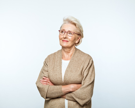 Portrait of confident senior woman standing with arms crossed, looking away and smiling. Studio shot, grey background.