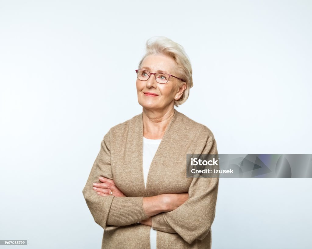 Portrait of smiling senior women Portrait of confident senior woman standing with arms crossed, looking away and smiling. Studio shot, grey background. Women Stock Photo