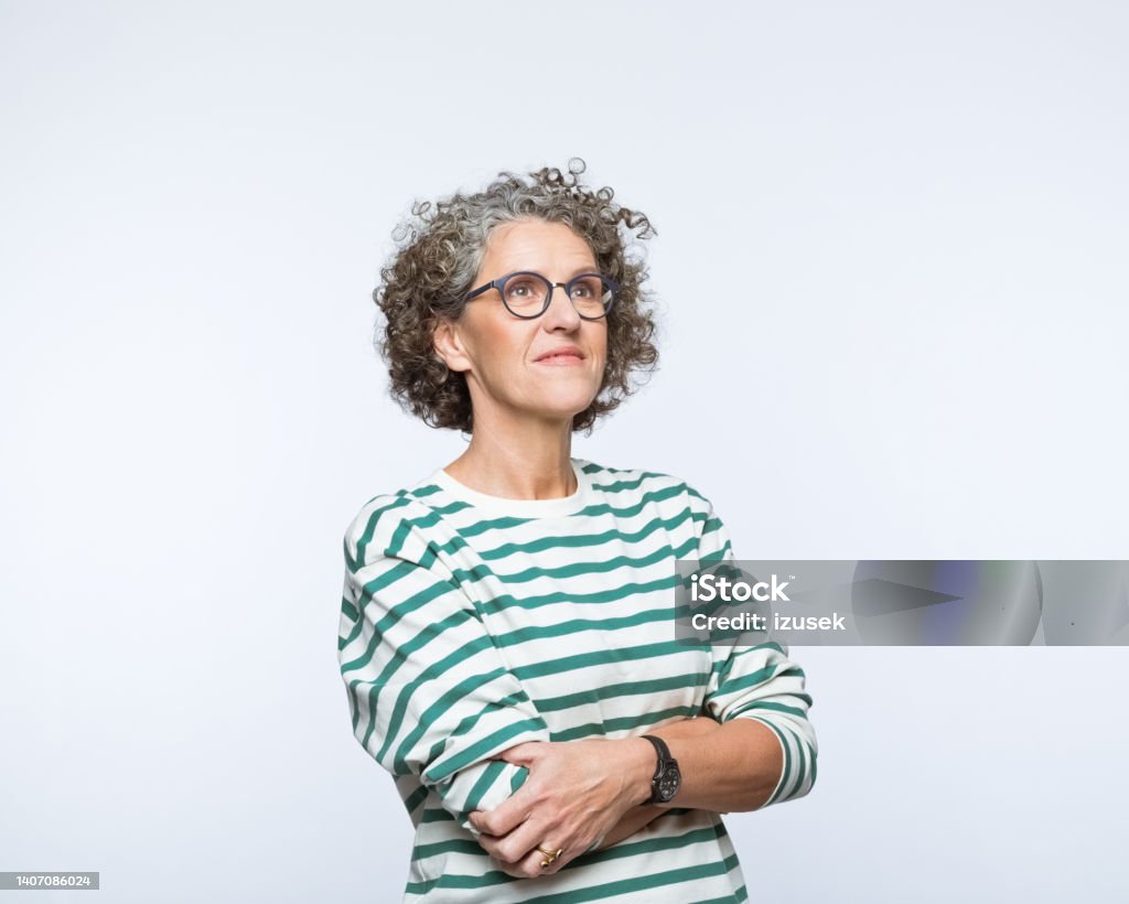 Portrait of thoughful mature women Portrait of mature woman wearing striped blouse and eyeglasses, standing with arms crossed and looking away. Studio shot, grey background. 50-59 Years Stock Photo
