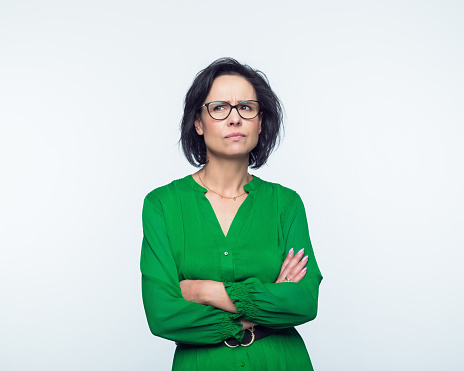 Portrait of confused mature woman wearing green dress, standing with arms crossed and looking away. Studio shot, grey background.