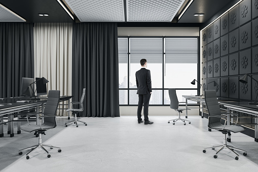 Thoughtful young businessman standing in modern dark concrete coworking office room interior with window, city view, curtains and furniture. Ceo and boss concept