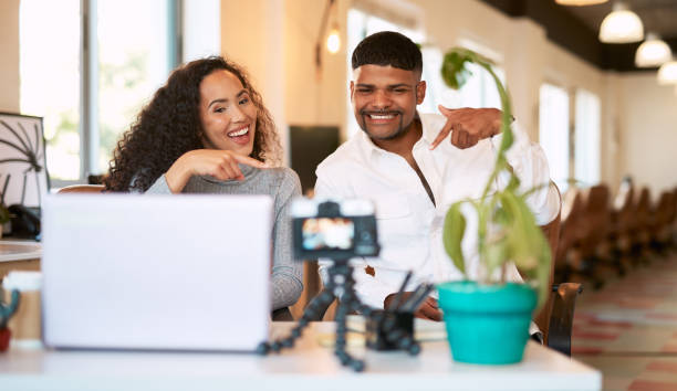 istock Young couple vlogging on digital camera, using laptop and sitting close together in home living room. Smiling happy boyfriend and girlfriend using hand gesture and pointing for social media followers 1407090657