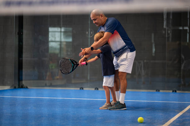 monitor enseñando clase de pádel a su niño, su alumno - entrenador enseña al niño pequeño a jugar al pádel en la pista de tenis cubierta - torneo de tenis fotografías e imágenes de stock