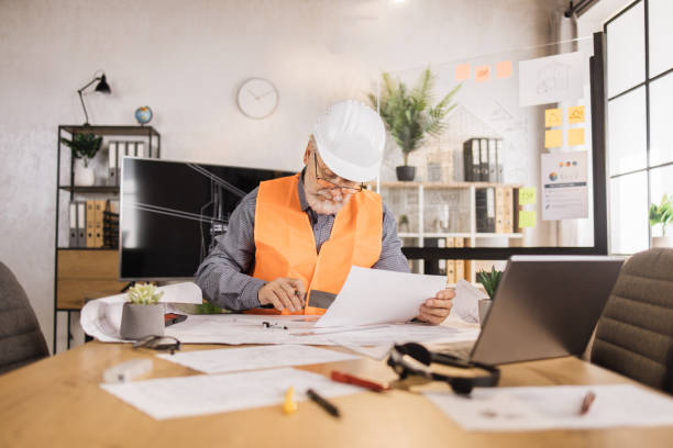 Senior builder, engineer in reflective vest uniform and hard hat on desk in office.