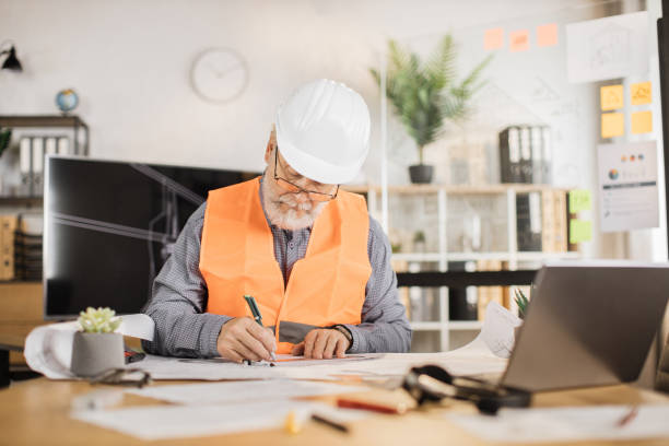 istock Senior builder, engineer in reflective vest uniform and hard hat on desk in office. 1407093413