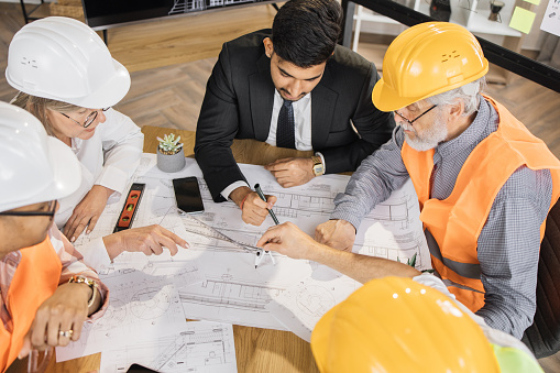 Qualified architects, engineers and designers having business meeting at office. Multicultural colleagues in suits and helmets sitting at table and looking at blueprints. Construction concept.