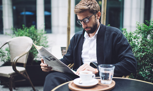 Serious bearded well dressed businessman in eyeglasses reading last news in newspaper while sitting with cup of coffee in street cafe