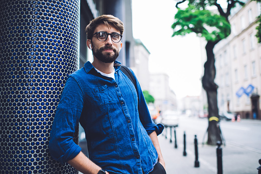 Young bearded male in casual outfit and eyeglasses standing on street in confident pose with hands in pockets and listening to music with wireless earphones