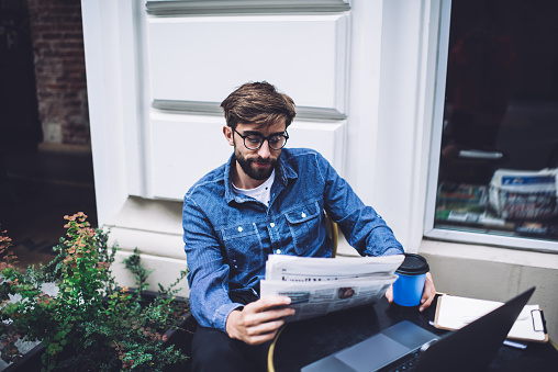 Male in eyeglasses and denim jacket sitting at black table in front of laptop while reading paper news and taking disposable cup of coffee in sunlight