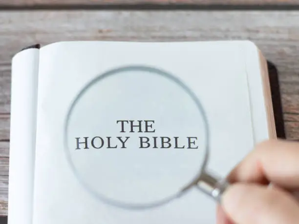 Human hand holds a magnifying glass on top of an open Holy Bible book on a wooden table. Search Scripture, biblical education, and seek God Jesus Christ concept. Top view. A closeup.