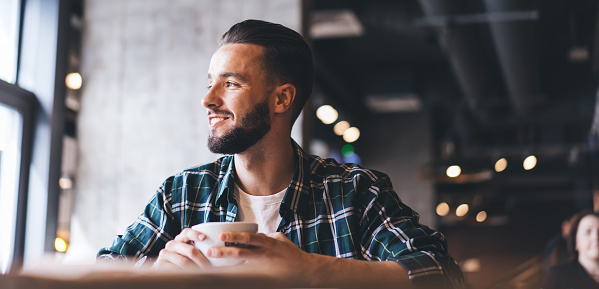 Positive bearded male client in checkered shirt drinking cup of coffee while resting at table in loft cafeteria and looking at window during break
