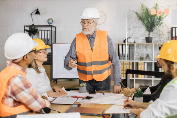 istock Multiracial architect people in helmets sitting at table with blueprints and modern devices. 1407098600