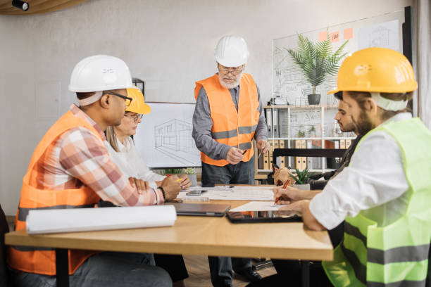 istock Multiracial architect people in helmets sitting at table with blueprints and modern devices. 1407098602