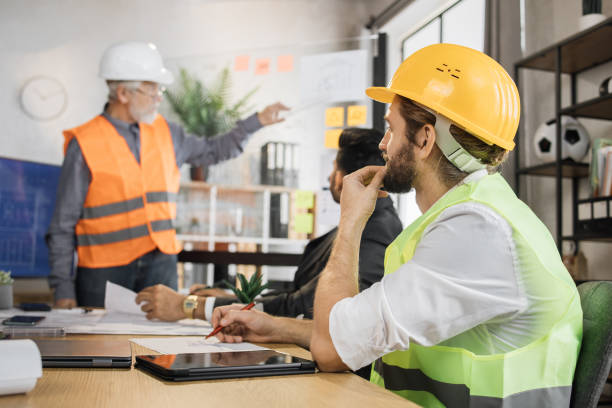 istock Attractive man in reflective vest sitting at table with males architects in uniform and helmets 1407098604