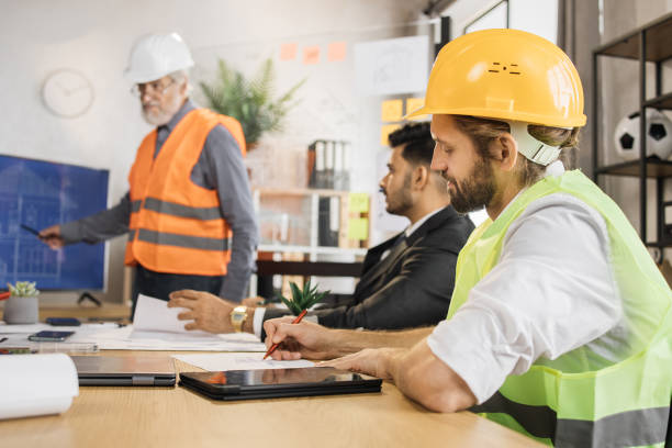istock Attractive man in reflective vest sitting at table with males architects in uniform and helmets 1407098606