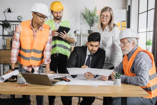 istock Multiracial architect people in helmets sitting at table with blueprints and modern devices. 1407098628