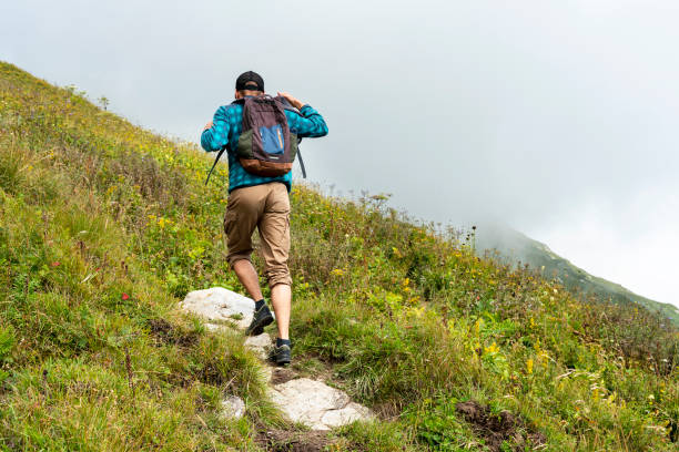 Rear view young man with a backpack walking uphill on a mountain path in fog among lush vegetation healthy and active lifestyle hiking outdoor activities, landscape