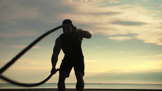 Bodybuilder training outdoors doing an exercise with battle ropes against the backdrop of sunrise. Training lifestyle.Slow Motion