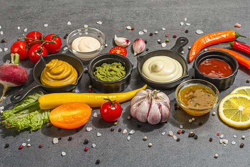 Set of sauces and fresh vegetables. Ketchup, mustard, mayonnaise, wasabi, sweet curry, and BBQ. Spices and herbs, trendy hard light, dark shadow. Stone concrete background, flat lay
