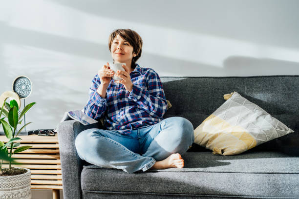 deep in thoughts, dreaming woman drinking coffee sitting on sofa in sunlight beams after reading a book at home in cozy modern interior with green plants. offline lifestyle. leisure and entertainment. - vehicle interior green sofa indoors imagens e fotografias de stock