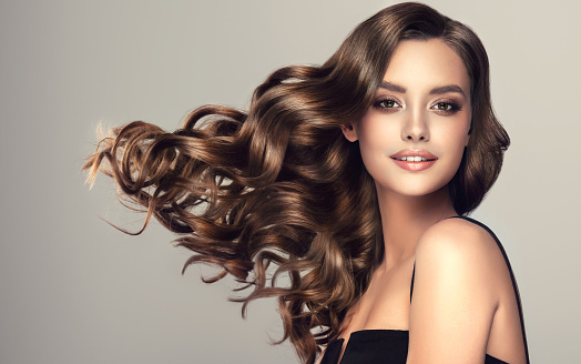 Beautiful woman face is surrounded by dense curls of long brown, well groomed hair. Model with long, wavy hairstyle and vivid makeup. Flying hair. Hairdressing art, beauty and style.