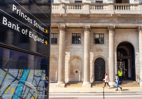 Close-up on a sign for the Bank of England, with pedestrians passing the institution's main entrance in the background.