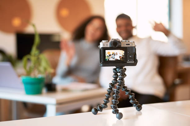 jovem casal vlogging em uma câmera digital enquanto sentados juntos em um sofá em uma sala de estar de casa. sorridente, namorado feliz e namorada usando gestos de mão e acenando para o canal de mídia social - digital video camera - fotografias e filmes do acervo