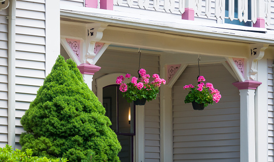 Twin hanging pots of pink geraniums suspended from the porch of a Cape Cod house.