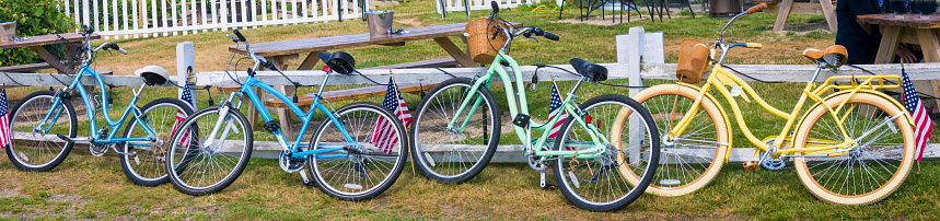 A row of four colorful beach bicycles lean against a fence surrounding an outdoor eating area of a Cape Cod restaurant.