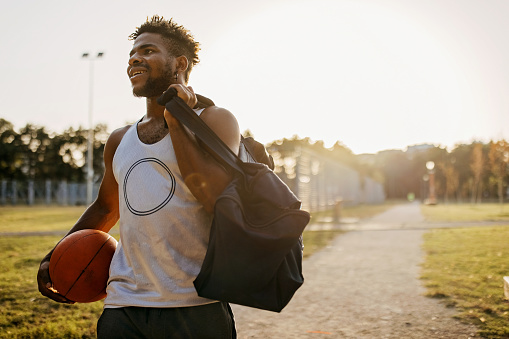A young African American man in sports clothes is holding a bag and a basketball, while walking down a footpath with a smile on his face.