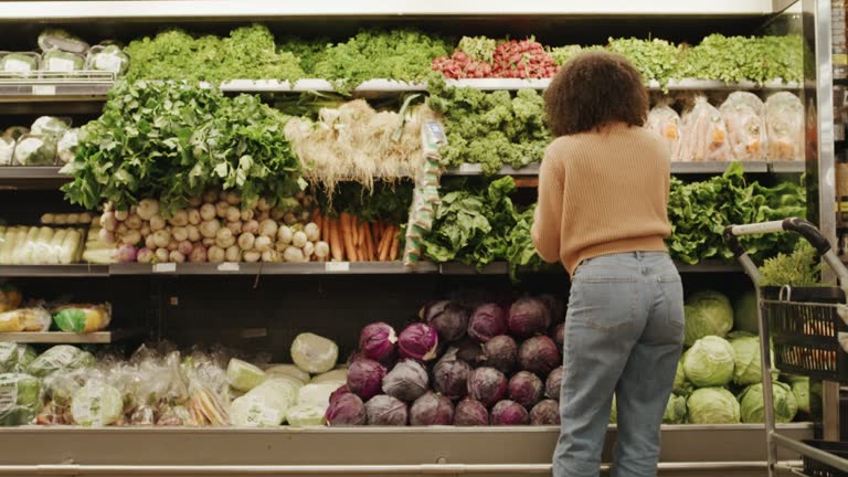 Rearview of a woman buying vegetables in a supermarket for diet and nutrition.Young female shopping for fresh produce in a local grocery store. Choosing leafy greens from a shelf to cook healthy
