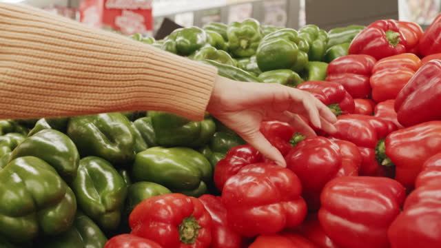 Closeup of one woman shopping for fresh fruit and veg produce on shelf in aisle of supermarket grocery store. Hands of customer choosing to buy red and green pepper to cook meal for healthy diet