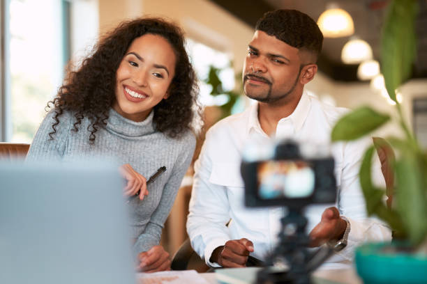 istock Young couple vlogging on digital camera, using laptop and sitting close together in home living room. Smiling happy boyfriend and girlfriend using hand gesture and pointing for social media followers 1407126773