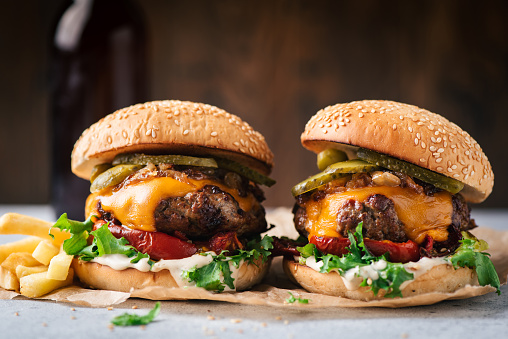 Two juicy craft cheeseburgers with roasted bell pepper and pickles served with french fries on parchment paper, wooden background