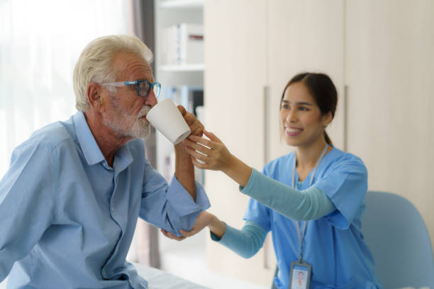 Asian elderly caregiver woman sitting on a hospital bed next to an older man helping drink a glass of water from the bedroom at home. stock photo