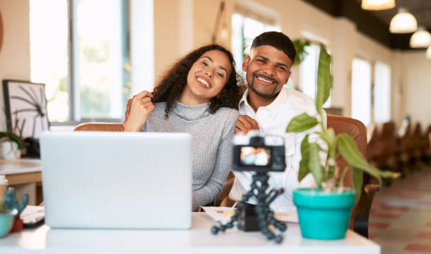 istock Young couple vlogging on tripod camera while sitting close together on sofa in a cafe. Smiling influencer boyfriend and girlfriend using laptop while working on content for social media followers 1407133257
