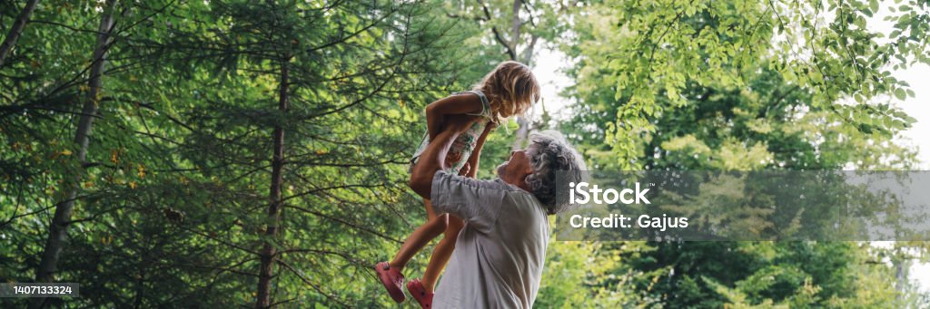 Wide view image of beautiful bond between a grandfather and a granddaughter Wide view image of beautiful bond between a grandfather and a granddaughter - grandpa lifting her grandchild high up in the air outside in green summer forest. Family Stock Photo