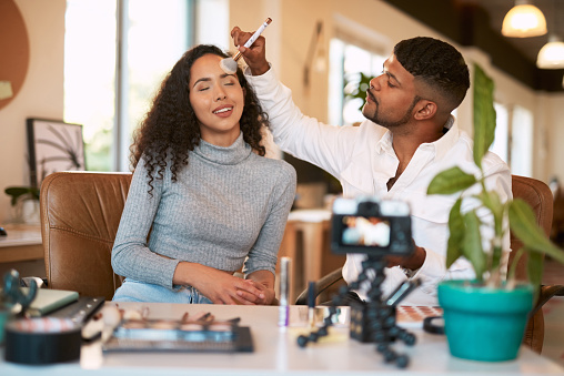 Male makeup artist applying cosmetics to female friends face. Queer beauty freelancer testing shades on partners skin. Influencer live streaming video while doing colleagues makeup in online tutorial