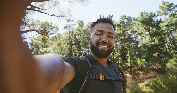 Active fit african american male taking a selfie photo and smiling while out on a hike in a natural green forest in the morning on a summer day. Healthy hiker enjoying the beauty of outdoor nature